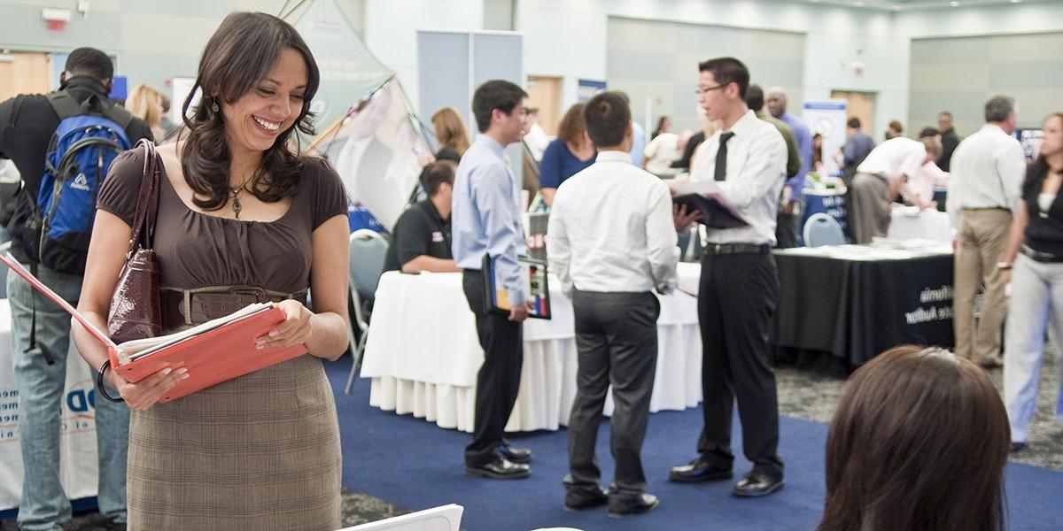 A student standing and smiling while holding an open binder and looking down at a person seated at a table. 